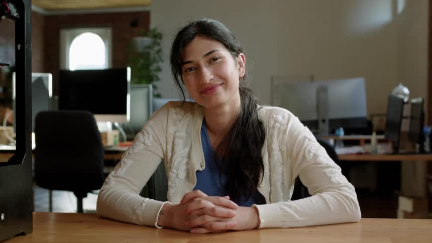 A woman is sitting at a desk with her hands clasped together and a smile on her face