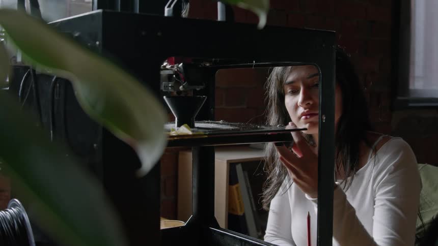 A woman observes a 3d printer creating one object while she holds another object in her hand