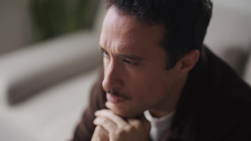 A man is sitting at the edge of the couch reaching moving the screen around on a laptop computer that is on the coffee table in front of him as he thinks about what he is doing he moves his hands together a rests them in front of his face