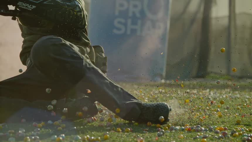 Paint ball competitor slides across ground strewn with paint balls near sign for paint ball manufacturer
