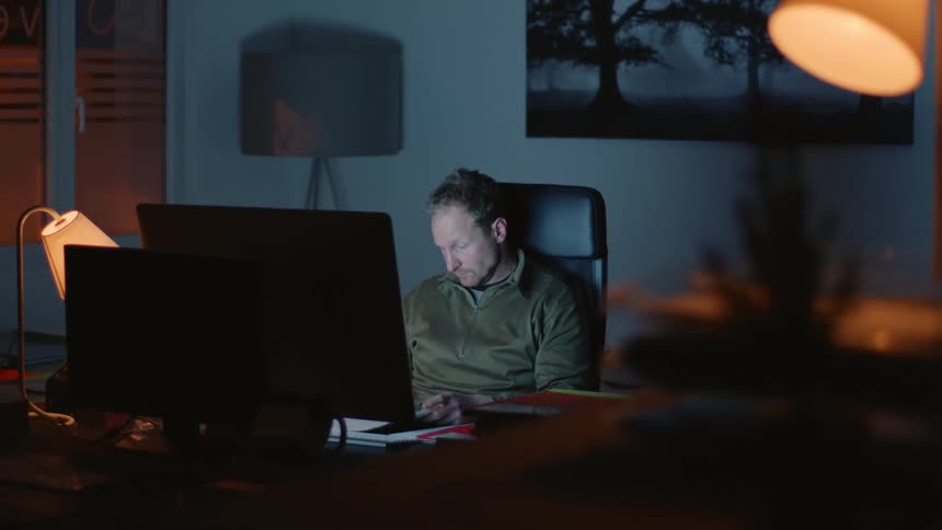 Late Night Office Work: Man Organizing Documents at Desk with Computer