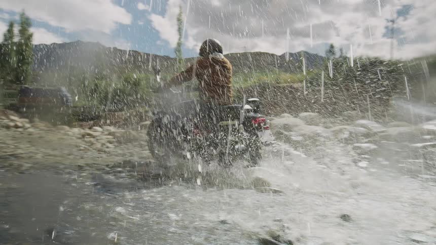 Man rides motorcycle through rock strewn creek splashing water as large commercial truck moves across bridge ahead