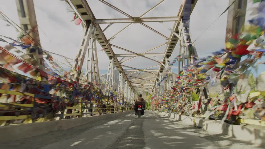 A person rides a motorcycle over a bridge covered in himalayan prayer flags