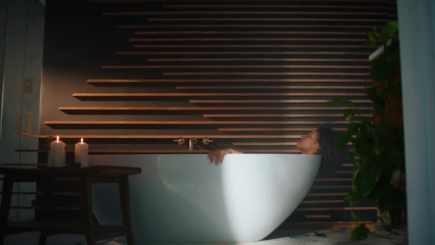 A woman lays in a bathtub relaxing with an abstract wall behind her and candles lit on a nearby table