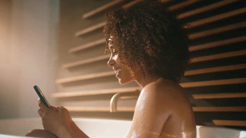 A woman is smiling and texting in the bathtub