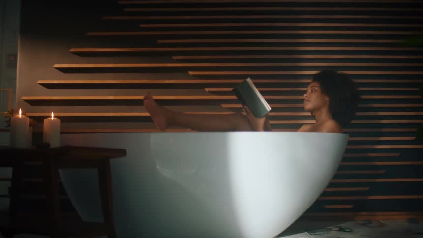 Woman relaxing in a white tub reading a book by candlelight