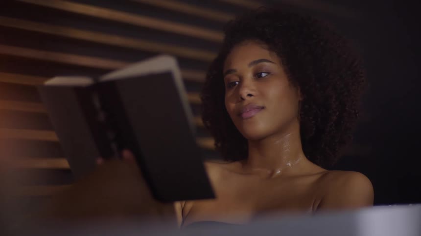 A beautiful black woman reads a book while soaking in a bathtub