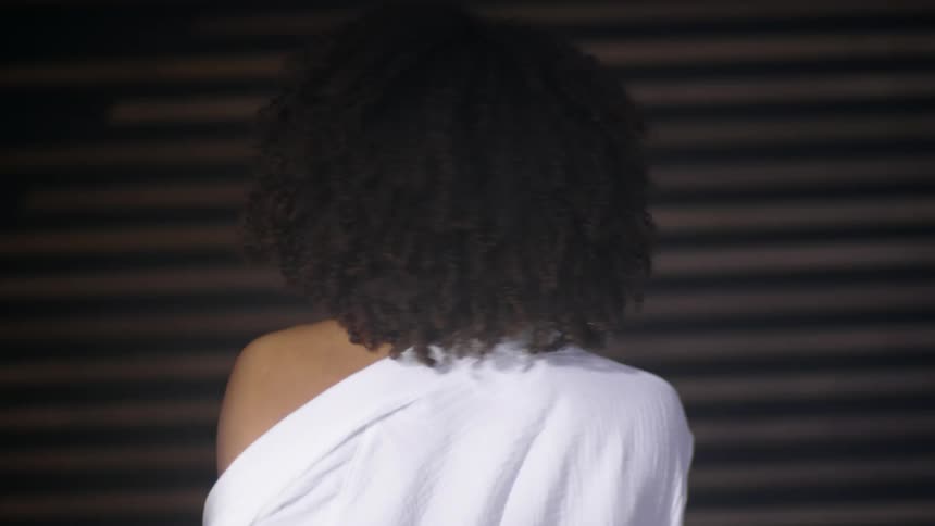 Black woman discarding white robe in front of a window shade