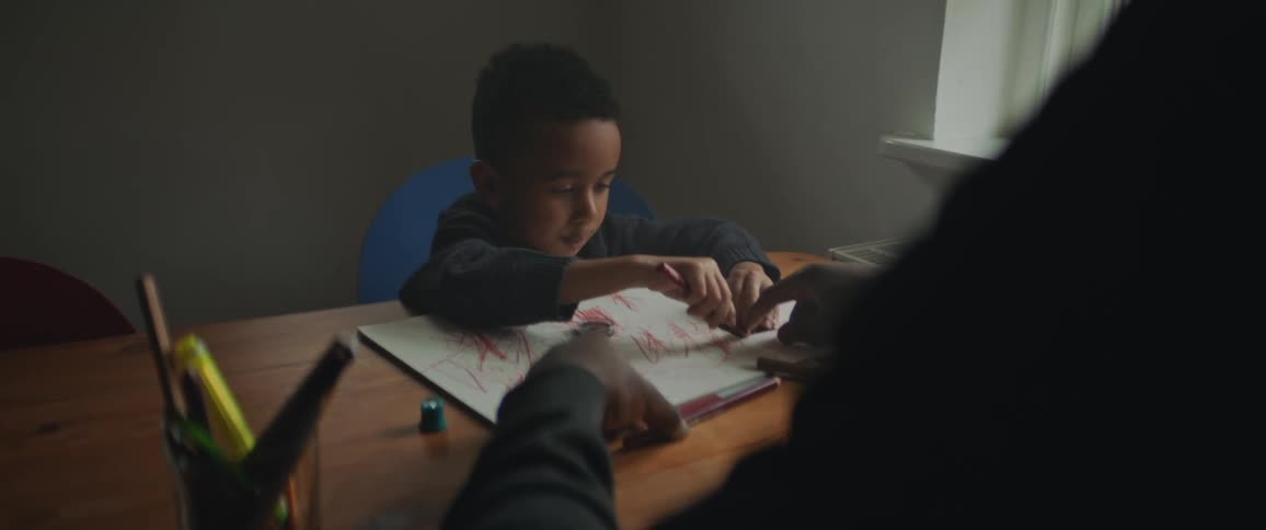 Parent sitting at table points at paper as son colors on it