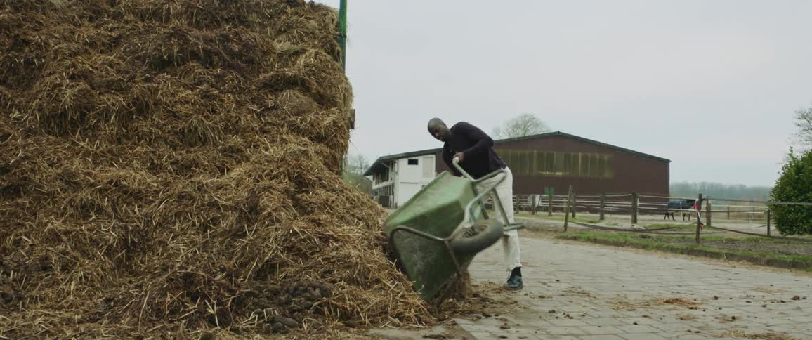 Farm worker pushing a wheelbarrow empties it on a pile of hay on the side of the road