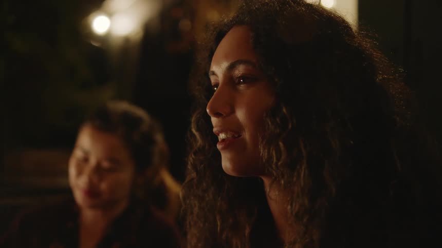 Two women with curly hair and ponytail sing a song smiling at night