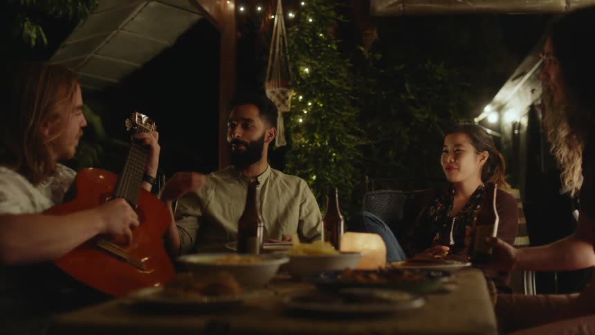 A group of friends sitting at desk talk to each other playing guitar and drinking beer