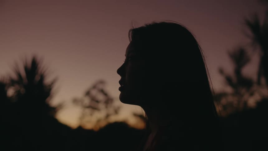 The silhouette of a womans side profile as she stands outside with trees in the distance behind her and a pink sky
