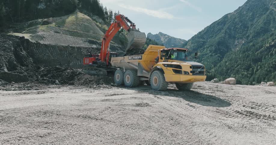Digger fills back of truck with dirt on plateau overlooking valley and conifer covered mountains