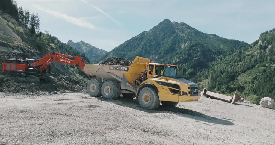 Man drives truck to haul dirt away from busy digger with forest covered mountains in the background