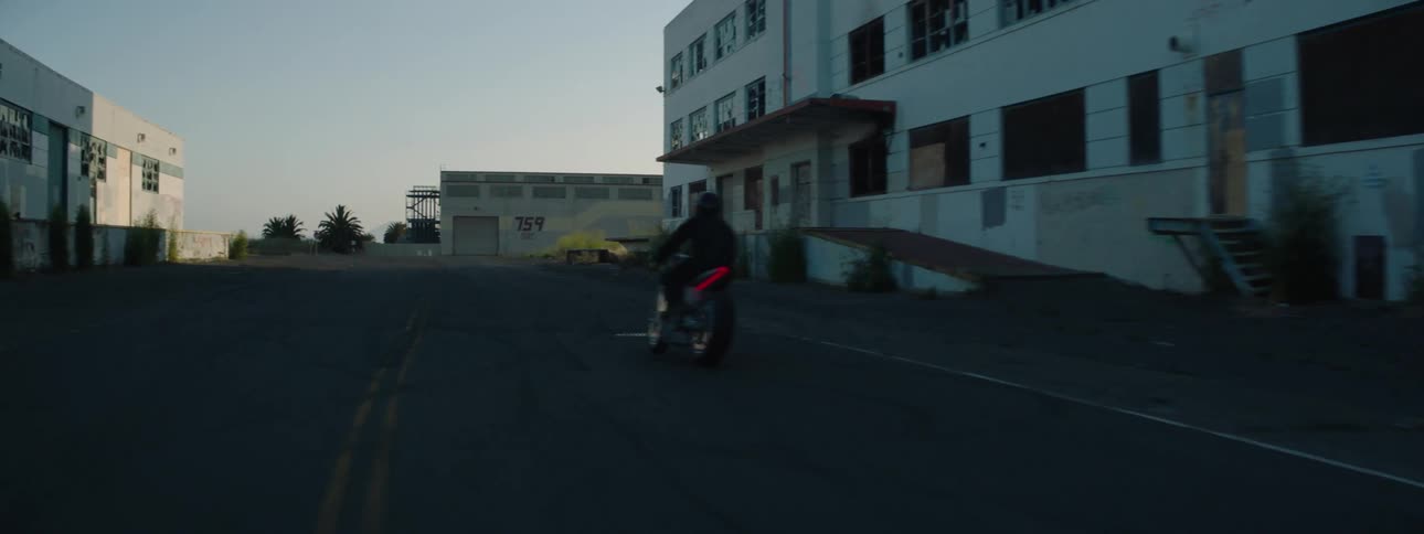 Motorcyclist rides along road past buildings on sunny day