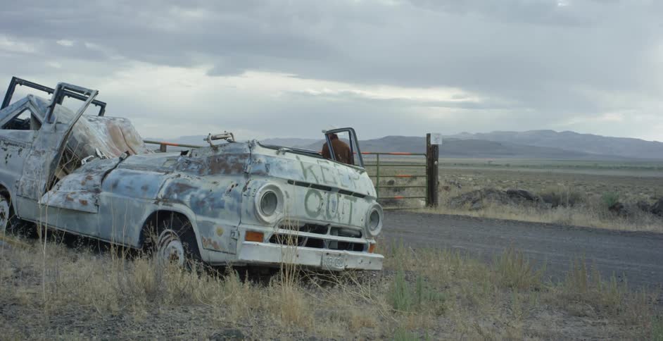 An old wrecked car on the side of a road as a man wearing a white cowboy hat walks by on the road