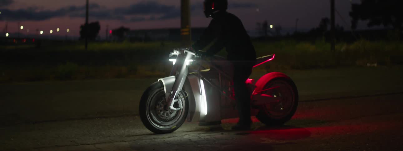 Man with helmet jacket and pants gets on bike riding off at sunset