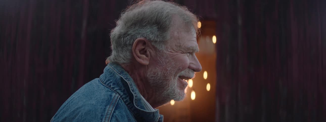 An elderly man is speaking and grinning as if remembering something bittersweet