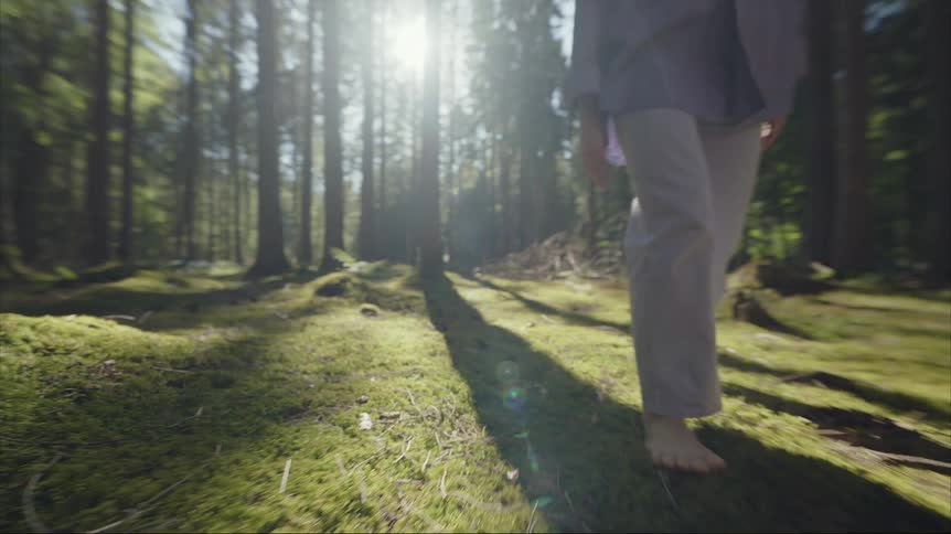 Woman with blue shirt and beige pants walks barefoot on green grass in the woods