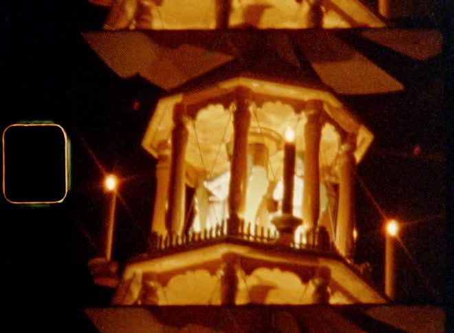 Tilting Super-8 film footage of lights glowing inside a Christmas tower or carousel with figures and sculptures of angels and Epiphany in the dark in front of the black night sky illuminated with burning candles as it spins or turns