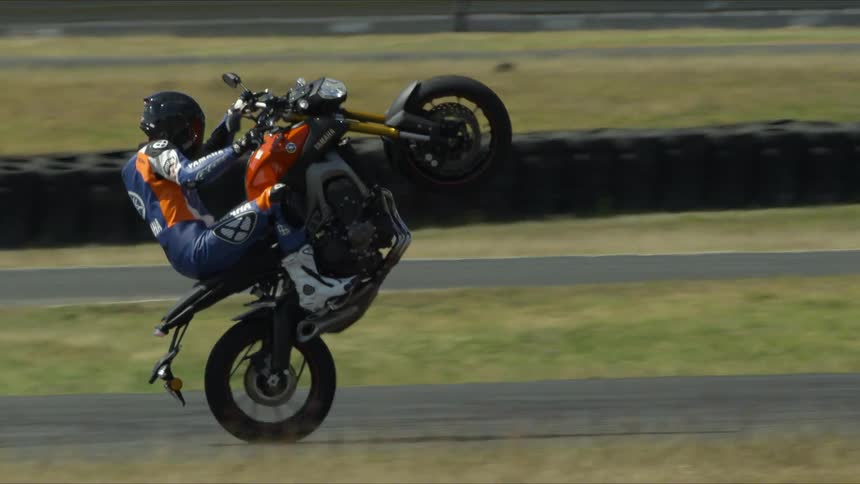 A stunt driver rides a motorcycle with the front tire in the air on an asphalt track