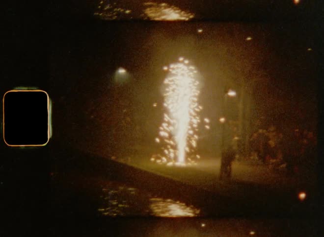 A group of people are standing near the coast watching fireworks shooting up as sparks glow in the darkness of night reflecting on the water