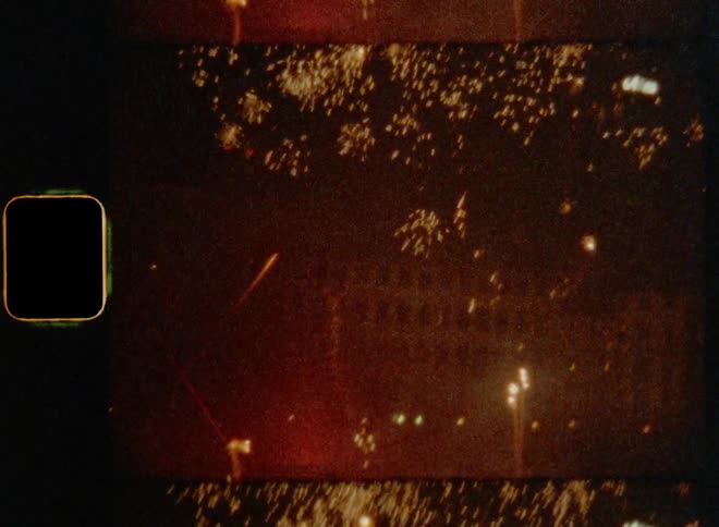 Old film footage of fireworks flying into the sky in front of a large building next to a waterfront during night time