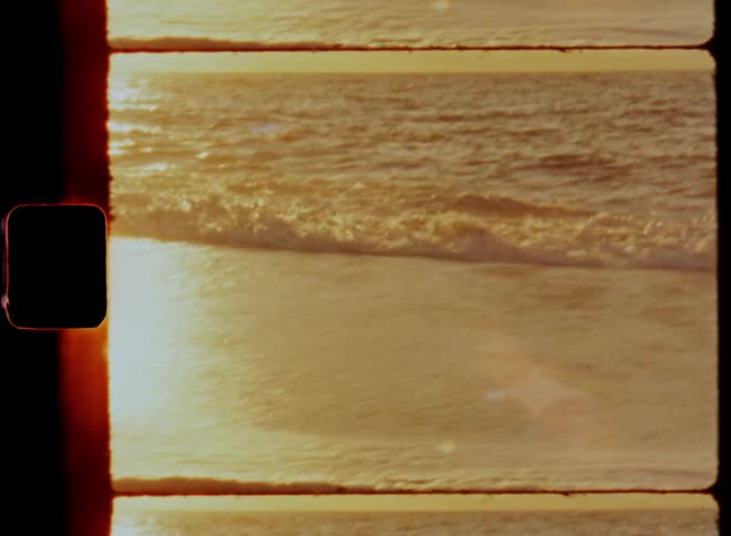 The ocean waves crashing up on the sandy beach as a yellow glow caused by the setting sun casts over the sea and sand on 8 mm film with lens flare and reflections