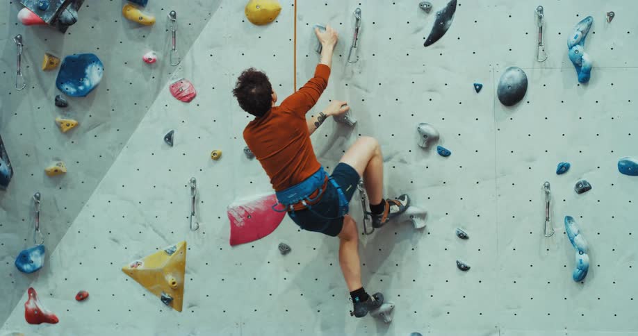 A teenage boy has a harness and rope around his waist as he scales a climbing wall and slips as he swings and hangs from his rope