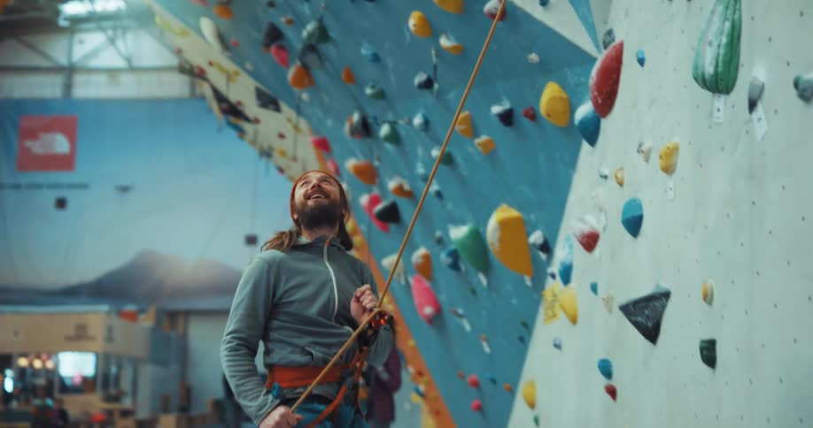 A man is standing near a climbing wall as he holds the rope attached to his harness and jumps on the side of the wall and laughs as he speaks to someone