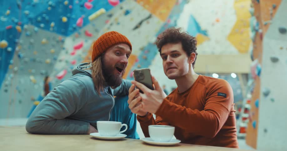 Two men sit and excitedly chat while drinking coffee and viewing a smart phone