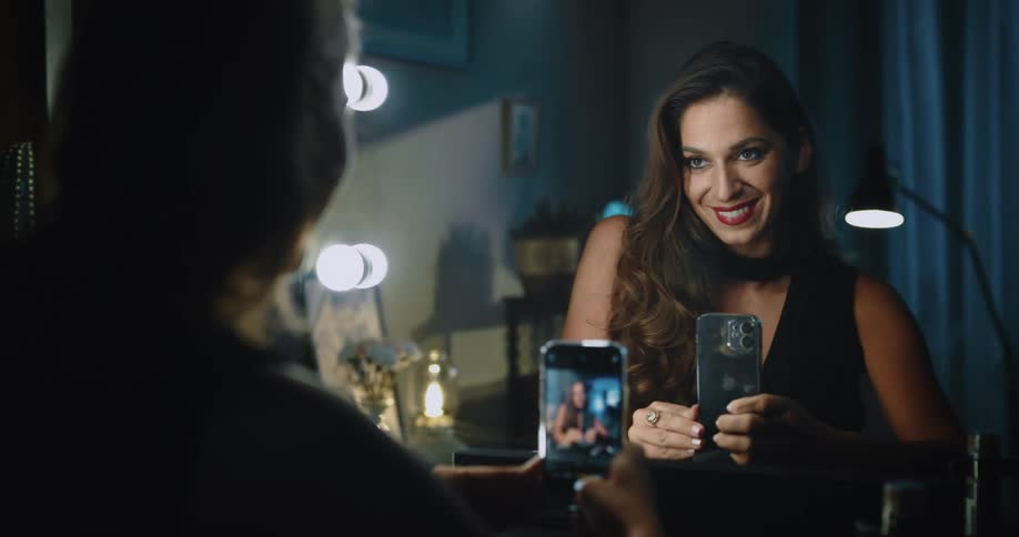 A woman sitting in front of a mirror grabbing her phone and taking selfies while smiling