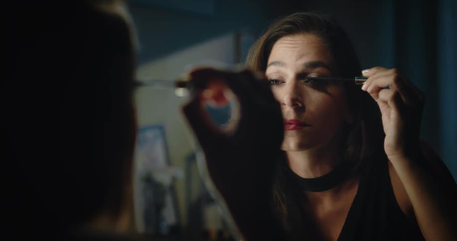 A woman is sitting in front of a mirror as she puts mascara on her eyelashes