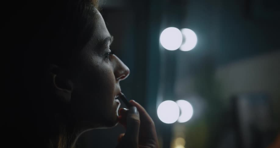 A woman is sitting in front of a vanity mirror and applying a lip pencil to her lips