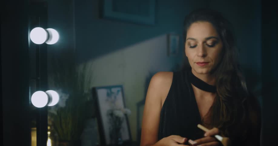 A woman walks into a room and sits down in front of a lighted mirror and looks at herself and rubs makeup on her finger under her eye