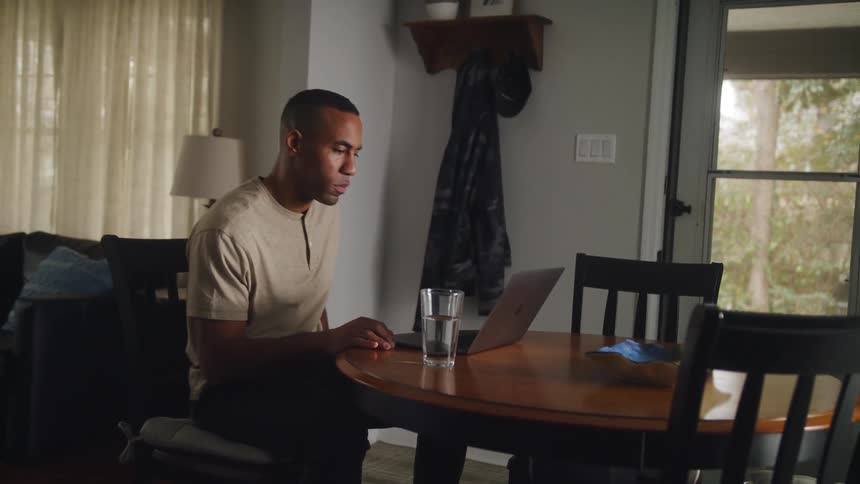 A man walks to a small table in his house and sits down to open his laptop
