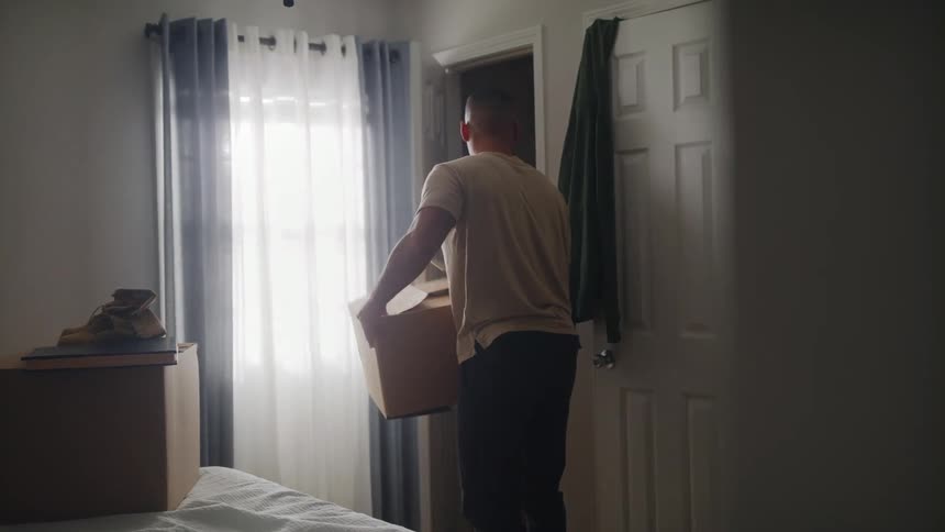 A man putting clothes into a cardboard box in a bedroom