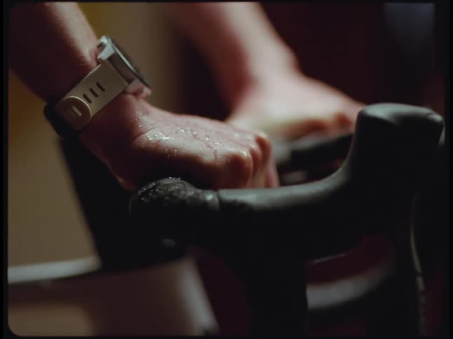 A womans sweat covered hands can be seen gripping a stationary bicycles handlebars