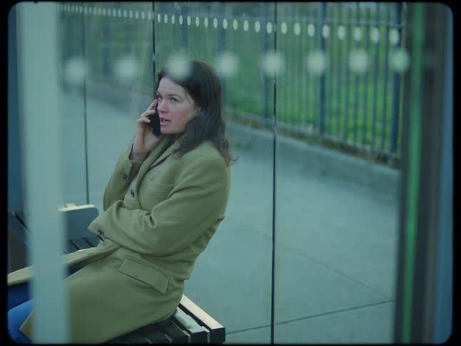 A woman wearing a long coat is sitting on a bench near a window as she talks on her phone