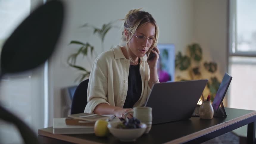 A woman is sitting at a desk working on a laptop computer as she talks on her phone