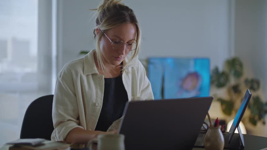 A woman is sitting at a desk with a laptop computer and a tablet as she types on the keyboard