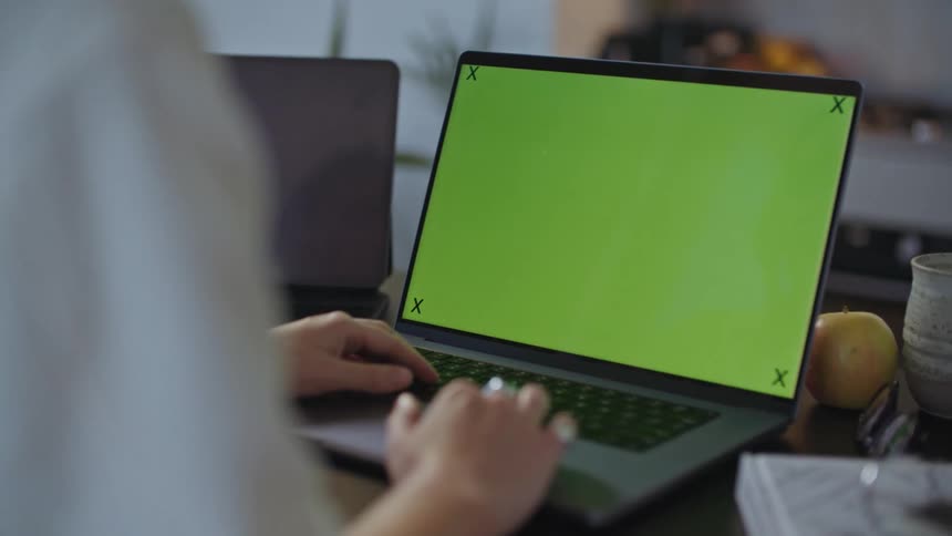 A woman is sitting at a table typing on a laptop computer with a green screen