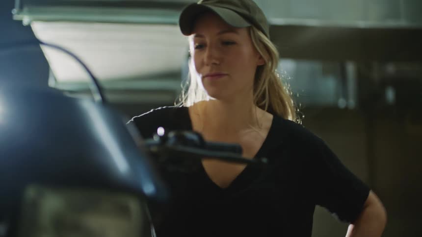 Blonde woman with dark green cap and black t shirt looks at dirt bike and smiles slightly