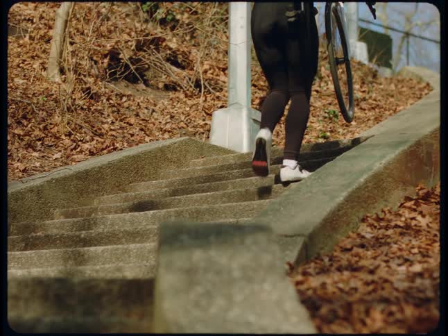 A woman carrying a bicycle up concrete steps wearing a white helmet and black suit