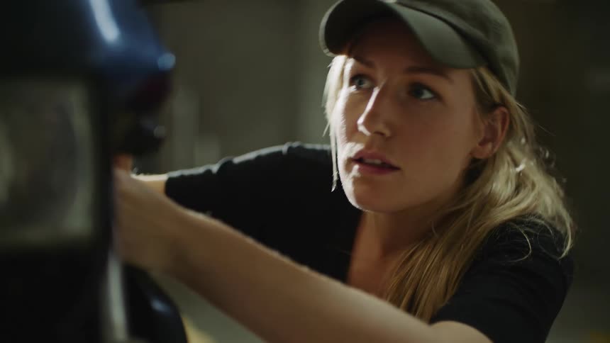 A woman with long blond hair wearing a cap is turning a ratchet as she works on a motorbike