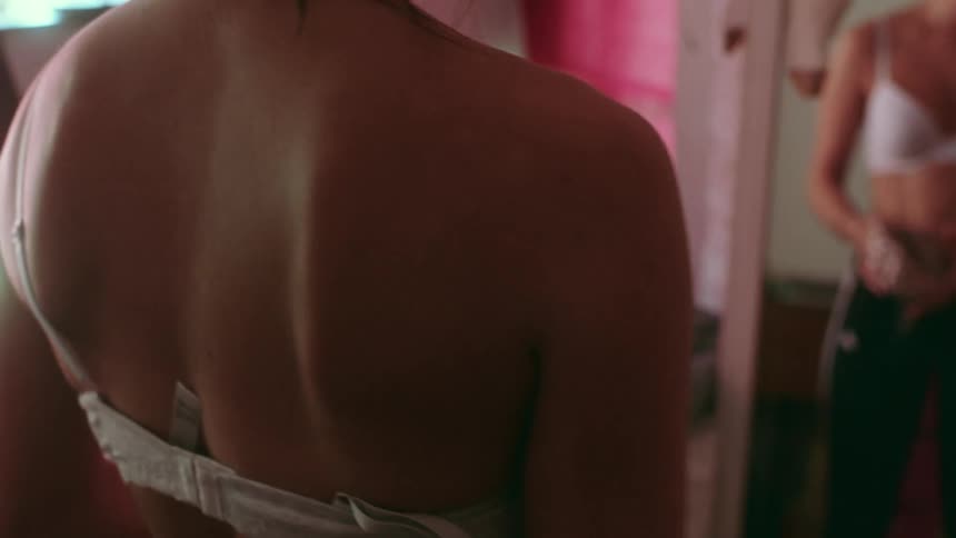 A young woman stands before a mirror and then starts to remove her bra