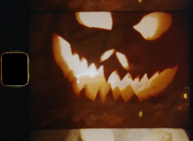 Old Super-8 film footage zooming out and revealing a scary Jack O'Lantern made of an orange pumpkin filled with a burning candle and smoke lit by a flickering flash light for Halloween celebration during a dark autumn night causing fear and stress
