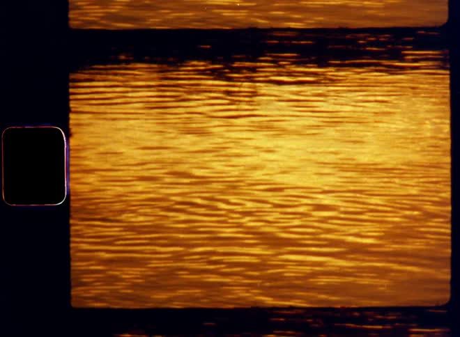 Super 8mm close up of the choppy water surface of a lake or river as a light breeze causes ripples and waves while the sun is setting during golden hour