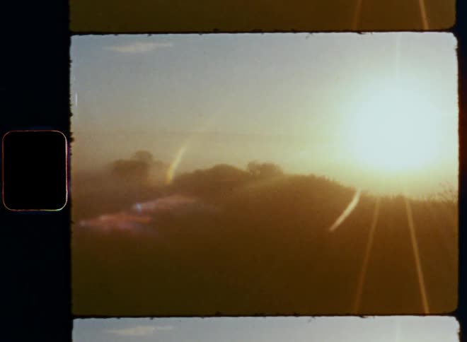 Panning Super 8mm wide angle footage of the rising sun shining brightly over a field with trees and vegetation under a blue morning sky on a clear day with fog and mist